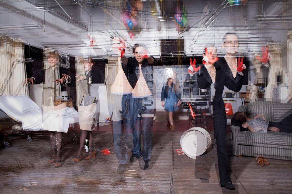 Grupa performerów w przestrzeni przypominającej salę szpitalną wykonuje ekspresyjne gesty. Na podłodze i w tle widać rekwizyty, łóżko oraz postać leżącą na kanapie.