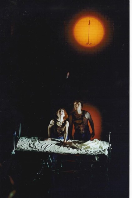 Kobieta i mężczyzna stoją za łóżkiem szpitalnym, oświetleni punktowym światłem na tle ciemnej sceny.