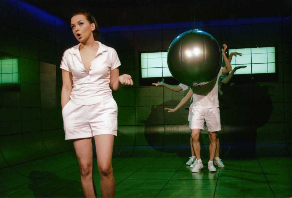 Na pierwszym planie aktorka w białym stroju wykonuje gest dłonią, jakby mówiła lub śpiewała. W tle dwie osoby niosą dużą kulę, częściowo zasłaniającą ich sylwetki. Zielona scenografia i kontrastowe światło nadają scenie performatywny charakter.