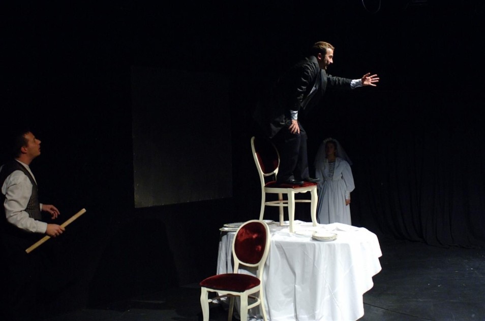Mężczyzna w czarnym stroju stoi na krześle ustawionym na stole przykrytym białym obrusem i wyciąga rękę przed siebie. Obok stoi inny mężczyzna z drewnianym kijem, w tle kobieta w sukni ślubnej.