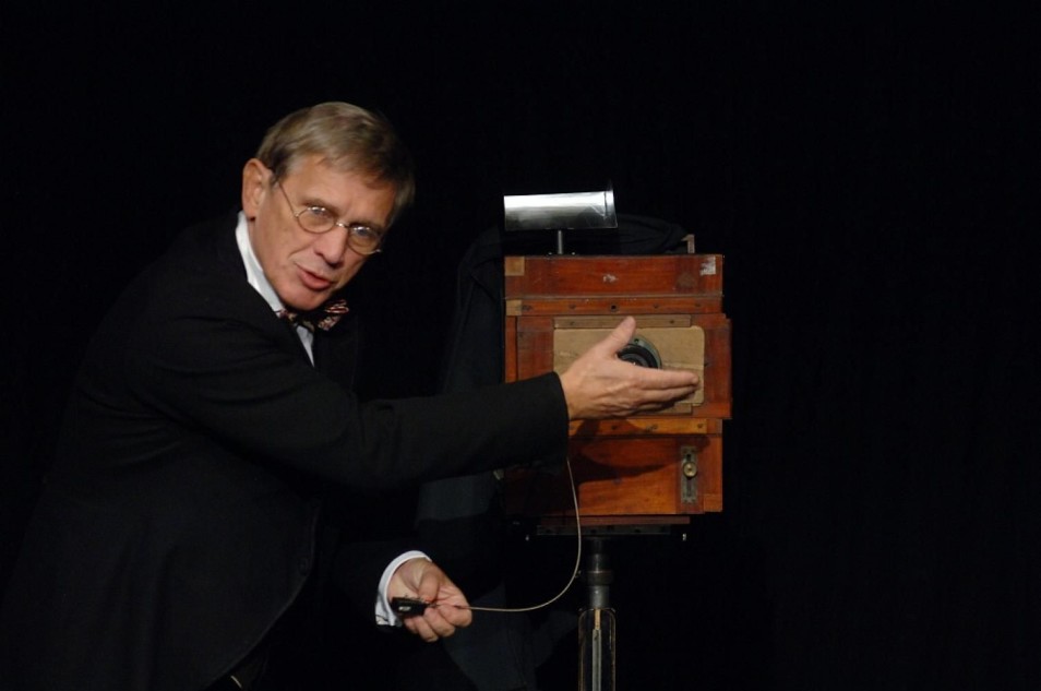 Mężczyzna w czarnym garniturze i okularach prezentuje stary aparat fotograficzny na statywie. Jedną ręką wskazuje obiektyw, w drugiej trzyma przewód. Tło sceny jest czarne i puste.