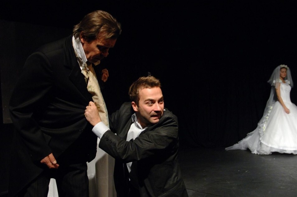 Dwóch mężczyzn w czarnych strojach znajduje się na pierwszym planie; jeden chwyta drugiego za klapę marynarki. W tle, po prawej stronie sceny, stoi kobieta w białej sukni ślubnej.