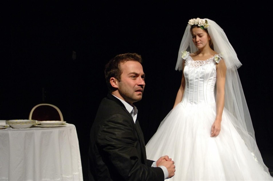 Mężczyzna w czarnym garniturze klęczy na scenie z wyrazem emocji na twarzy. Przed nim stoi kobieta w białej sukni ślubnej i welonie, patrząca na niego z góry. Obok widoczny stół z białym obrusem i talerzami.