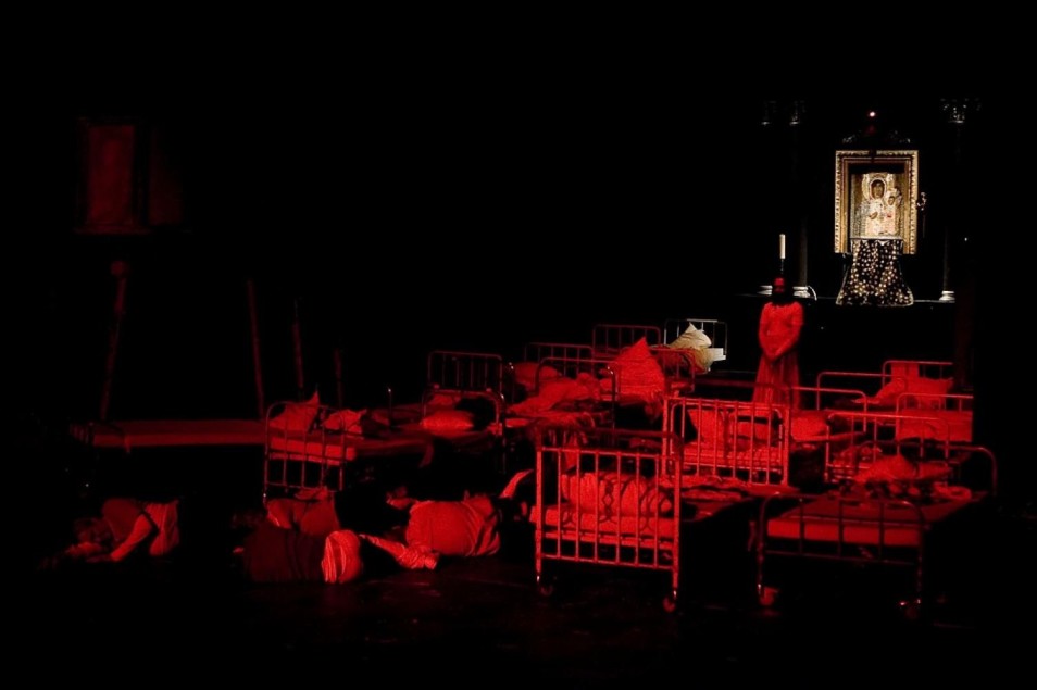 Mroczna scena oświetlona czerwonym światłem. Łóżka szpitalne wypełniają przestrzeń, część postaci leży nieruchomo. Z tyłu ikona religijna, atmosfera zagrożenia i niepokoju.