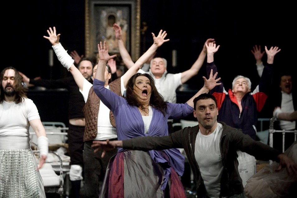 Zespół aktorów z uniesionymi rękami wykonuje dynamiczny, zbiorowy gest. W centrum kobieta z otwartymi ustami, jakby krzyczała lub śpiewała. Tło stanowią łóżka szpitalne i ikona.