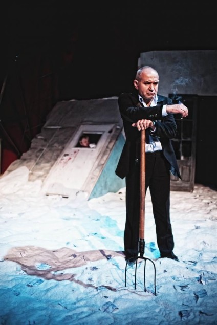 Starszy mężczyzna w garniturze stoi na zaśnieżonej scenie, oparty o widły. W tle widać zniszczone elementy scenografii i dym.