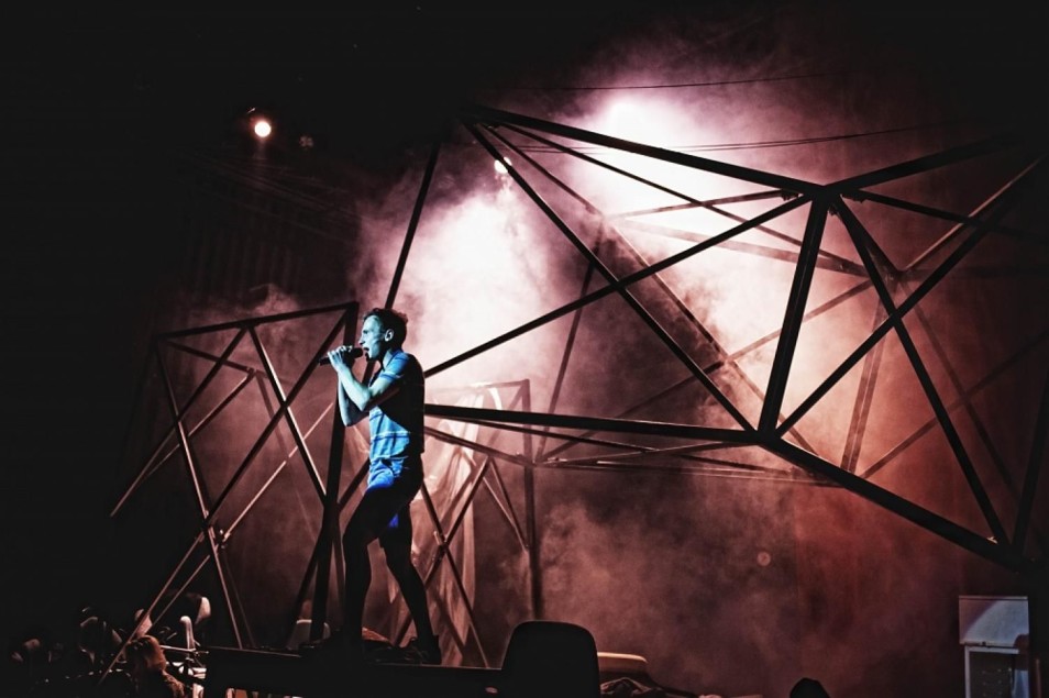 Performer stoi na podwyższeniu i śpiewa do mikrofonu. Za nim widoczna jest duża metalowa konstrukcja i gęsty dym, oświetlone teatralnym światłem.