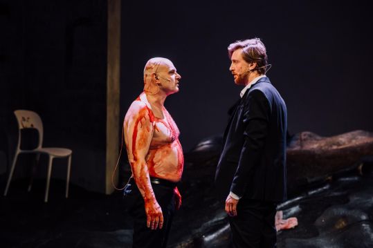 Zakrwawiony, półnagi mężczyzna (Robert Ninkiewicz) stoi twarzą w twarz z innym mężczyzną w garniturze (Piotr Biedroń); obaj patrzą na siebie w napięciu.