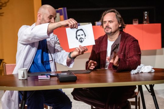 Dwóch mężczyzn siedzi przy stole. Lekarz (Piotr Chys) kartkę z rysunkiem twarzy, Pana Baran (Piotr Łukawski) przygląda się rysunkowi. Na stole stoją kubki i drobne przedmioty.