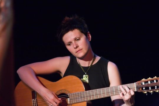 Dziewczyna z krótkimi włosami gra na gitarze. Ma czarną koszulkę bez rękawów i duży naszyjnik. Patrzy z natchnieniem w dół.