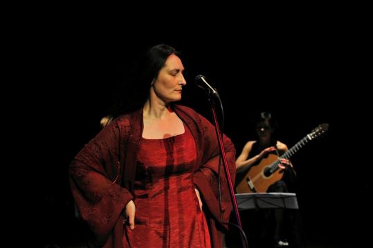 Marzena Nieczuja Urbańska w natchnieniu, z przymkniętymi oczami. Ręce na biodrach, przed nią mikrofon. W tle gitarzystka.