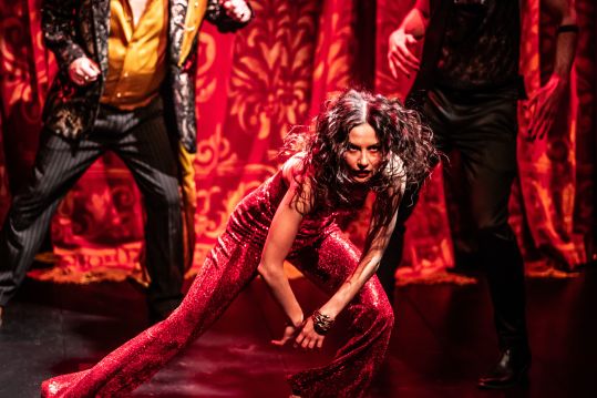 Zbliżenie na postać Izy (Agata Woźnicka). Nogi ugięte, jedna w wykroku w bok, ręce i tułów opuszczony, patrzy przed siebie. Ma rozwiane, ciemne, kręcone włosy a na sobie połyskujący czerwony kostium. W tle za nią dwie postaci i czerwona kurtyna.