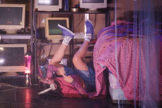 Laura w swoim pokoju. Leży na plecach obok łóżka z nogami w górze. Na głowie duże, białe słuchawki. Ma fioletowe Vansy i jasne, długie skarpetki w zebry. W tle monitory.