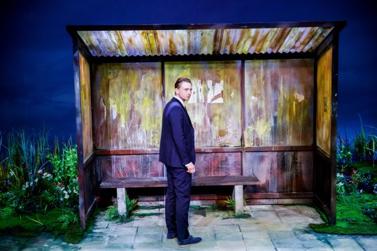 Mężczyzna w garniturze (Piotr Biedroń) stoi bokiem przed wiatą przystankową.