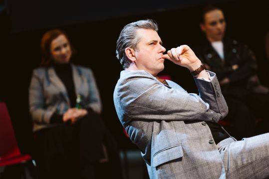 Profil mężczyzny w jasnoszarym garniturze (Maciej Konopiński). Dłoń przy ustach, wyraz twarzy skupiony, pokerowy. W tle sylwetki dwóch kobiet.