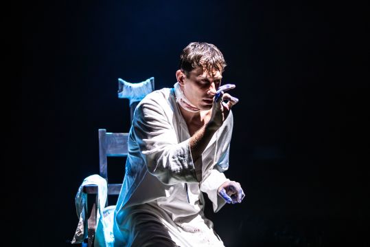 Mężczyzna w spitalnym stroju (Marcin Miodek) siedzi na krawędzi krzesła, w skupieniu podnosi palce jakby pisał w powietrzu, w chłodnym, niebieskim świetle na ciemnym tle.