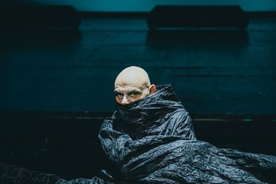 Na zdjęciu pojedyncza osoba (Robert Ninkiewicz) na scenie, owinięta ciemnym materiałem, z widoczną głową. Postać siedzi lub klęczy. Tło jednolite, niebiesko oświetlone.