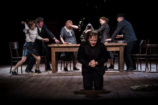 Scena teatralna: kilka osób w gwałtownym konflikcie wokół drewnianego stołu. Jedna postać klęczy na pierwszym planie, reszta gestykuluje i rzuca przedmiotami. Dynamiczna, dramatyczna sytuacja na pustej scenie.