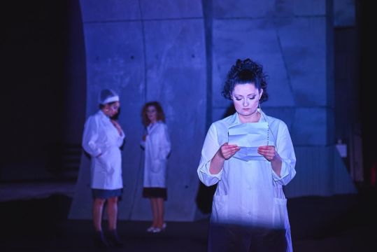 Kobieta w białym fartuchu stoi na pierwszym planie i czyta kartkę papieru. W tle dwie inne kobiety w podobnych strojach. Scena oświetlona chłodnym, niebieskim światłem.