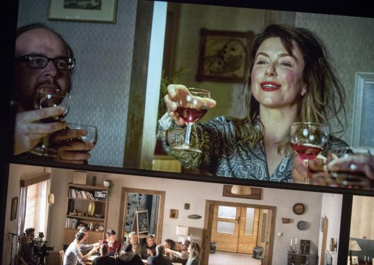 Grupa dorosłych siedzi przy stole w mieszkaniu i wznosi toast kieliszkami wina; ujęcie pokazuje jednocześnie scenografię i scenę z udziałem aktorów.