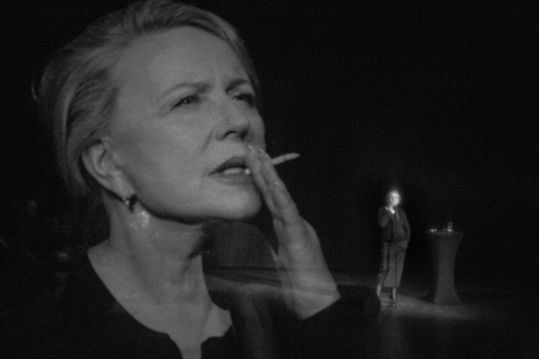 Zbliżenie twarzy kobiety palącej papierosa; w tle, na scenie, ta sama postać stoi przy mikrofonie w ciemnej przestrzeni.