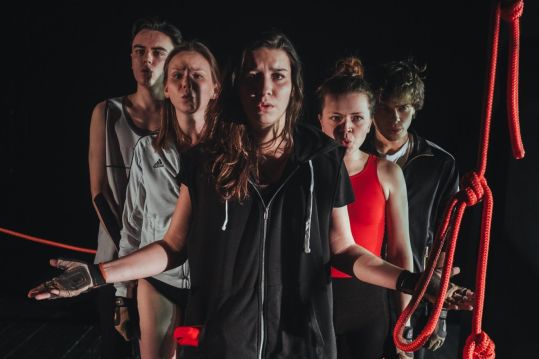 Pięcioro młodych aktorów stoi w szeregu na ciemnej scenie, z napiętymi twarzami, kobieta na przodzie stoi z rozłożonymi rękami. Obok nich zwisają czerwone liny.