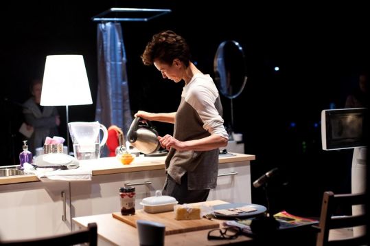 Aktorka nalewa gorącą wodę z czajnika do szklanego naczynia na kuchennym blacie scenografii.