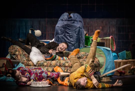 Trójka aktorów leży i siedzi na kanapie oraz podłodze wśród kolorowych rekwizytów; jeden z mężczyzn trzyma szklankę, scena wygląda na chaotyczną.