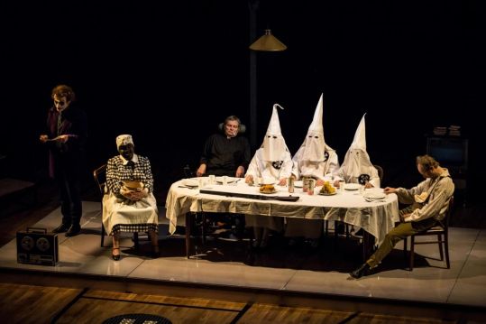 Przy długim stole na scenie siedzi grupa aktorów, część w białych kapturach i kostiumach; obok stoją inne postacie, a na stole znajdują się naczynia i jedzenie.
