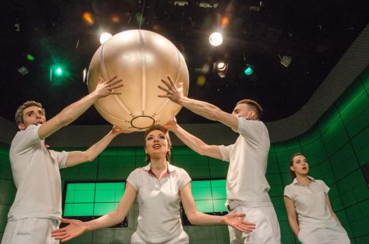 Na scenie czworo aktorów w białych strojach, dwójka unosi nad głową dużą, kulistą formę. Centralna postać stoi pod obiektem z rozłożonymi rękami i uniesioną głową. Zakrzywiona scenografia i punktowe światło nadają scenie choreograficzny charakter.