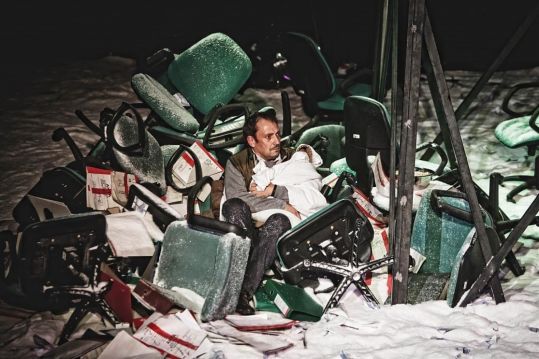 Mężczyzna siedzi pośród sterty przewróconych biurowych krzeseł i papierów, trzymając zawinięte niemowlę. Scena wygląda jak po katastrofie.