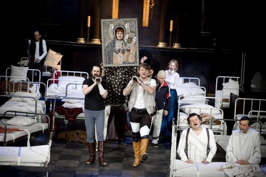 Grupa aktorów maszeruje między łóżkami szpitalnymi, niosąc ikonę Matki Boskiej. Postacie w różnych pozach, scena pełna napięcia i ruchu.