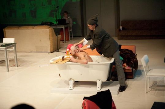 Kobieta pochyla się nad mężczyzną leżącym w wannie i podtrzymuje jego głowę. W tle widoczna kanapa, stolik i osoba pracująca przy komputerze.