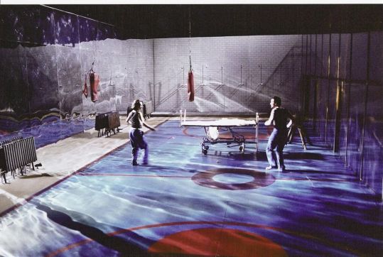 Dwóch aktorów porusza się po scenie stylizowanej na salę gimnastyczną z łóżkiem szpitalnym i wiszącymi workami.