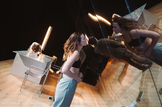 Aktorka stoi bardzo blisko szklanej ściany i patrzy na swoje odbicie; pozycja ciała i światło tworzą wrażenie konfrontacji i wewnętrznego napięcia.