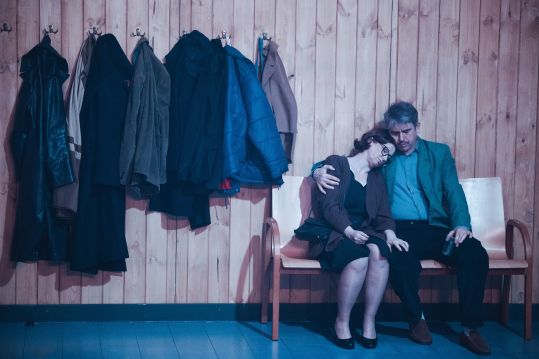 Mężczyzna obejmuje kobietę siedzącą obok niego na ławce; za nimi wiszą płaszcze na wieszakach.