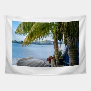 Tropical garden frames view over white clinker dinghy to bay with stand-up paddle-boarder in distance. Tapestry