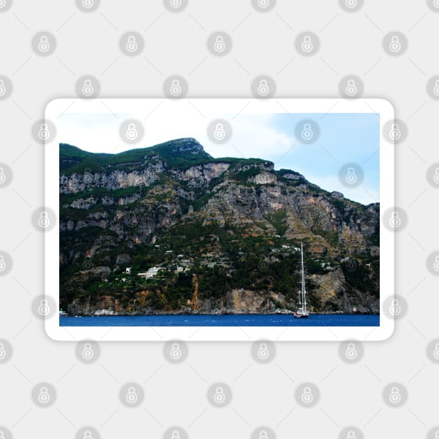 View in Amalfi coast at a huge mountain with rocky parts, greenery and buildings with a white boat underneath Magnet by KristinaDrozd