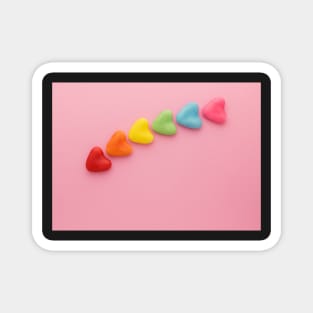 A variety of colorful candy hearts, in a rainbow shape, on a light pink background. Magnet