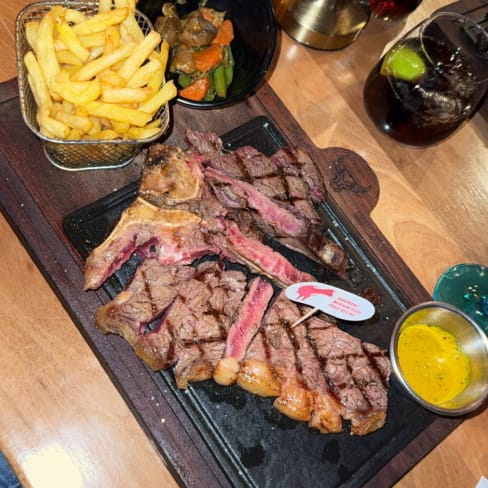 Une généreuse portion de faux-filet grillé, tranché pour mettre en valeur son intérieur juteux et tendre, est le plat principal. Accompagné de frites dorées et croustillantes, ainsi que de légumes mélangés, dont des carottes et des haricots verts, le tout est rehaussé par un bol de sauce moutarde. Présentation évoquant un repas copieux et satisfaisant.