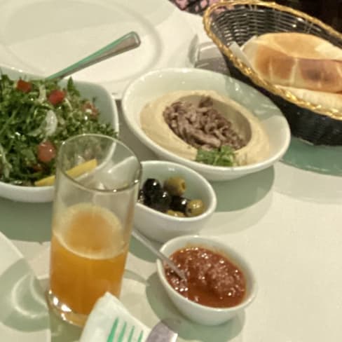A spread of Middle Eastern dishes featuring a fresh salad, creamy hummus with olive oil, olives, a chunky tomato sauce, flaky pastries, warm bread, and a refreshing orange beverage.