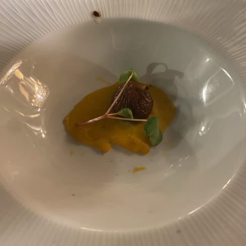 Purê laranja cremoso, possivelmente de abóbora ou cenoura, com um elemento de chocolate escuro no centro, guarnecido com microverdes, destacando a harmonia entre os sabores doces e salgados.