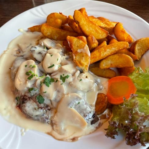 Zarte Fleischstücke, wahrscheinlich Rind, in einer cremigen weißen Sauce mit Pilzen, garniert mit frischen Kräutern, serviert mit goldbraunen Kartoffelspalten und frischem Salat sowie Karottenscheiben.
