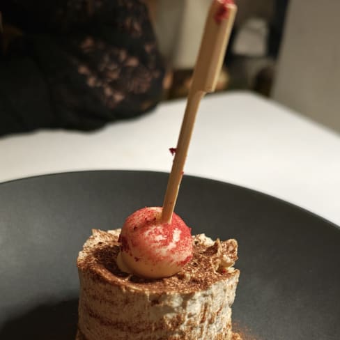 Zartes Schichtdessert in zylindrischer Form, wahrscheinlich Tiramisu, mit Kakaopulver bestäubt und einem glänzenden roten Element, das die Präsentation verfeinert.