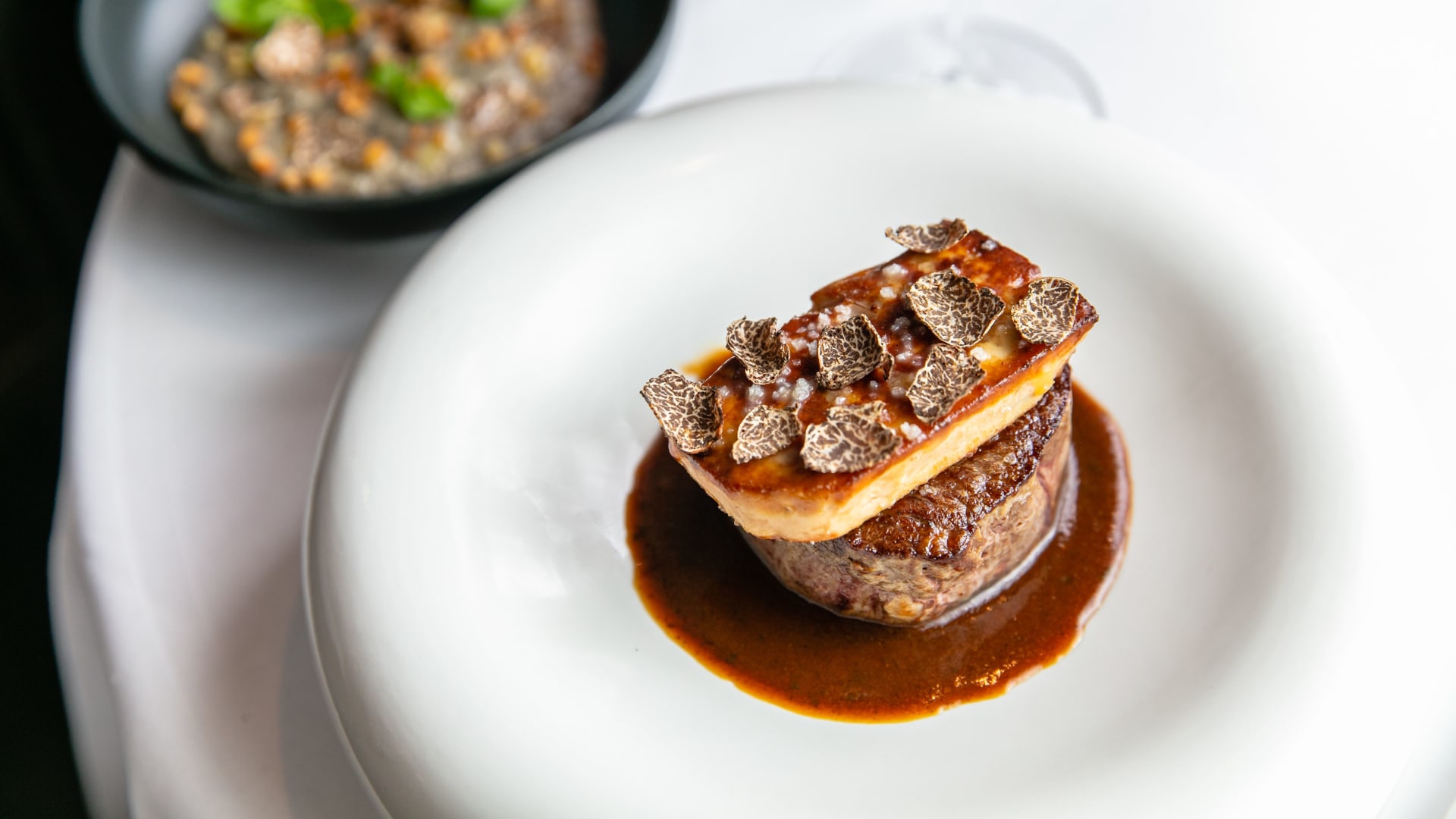Plat de viande de boeuf parfaitement saisie, surmontée de foie gras caramélisé, servie avec une sauce sombre et garnie de truffe, accompagnée d'un accompagnement de grains.