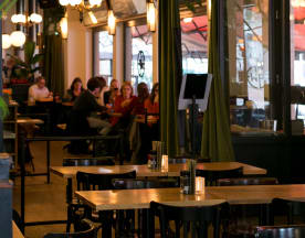 Het interieur van het restaurant is gezellig en warm, met houten tafels en stoelen die een uitnodigende sfeer creëren. Zachte verlichting van hangende lampen zorgt voor een ontspannen ambiance. Achtergrondgasten genieten van hun maaltijden, wat bijdraagt aan de levendige en comfortabele setting. Grote ramen laten natuurlijk licht binnen, terwijl groene gordijnen kleur en warmte aan de inrichting toevoegen. Een ideale plek voor informele diners en sociale bijeenkomsten.