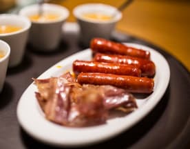 Salchichas ahumadas acompañadas de finas lonchas de carne curada y una salsa dorada, creando una experiencia de sabores y texturas.