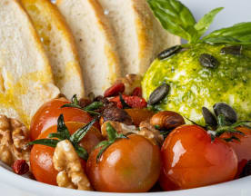 Queijo cremoso com ervas, servido em pesto verde, acompanhado de tomates cherry assados, nozes, bagas de goji e sementes de abóbora, com fatias de pão tostado.