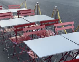 Espace de restauration en plein air avec des tables simples en surface grise et des chaises en bois rouge, offrant une atmosphère décontractée et accueillante.