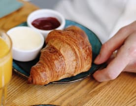 Versgebakken croissant, goudbruin en luchtig, geserveerd met romige boter en rijke rode jam, vergezeld van een glas sinaasappelsap.
